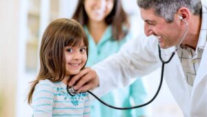 Family doctor examining young girl with stethoscope while mother watches; child looking at the camera. Family health center providing pediatric care